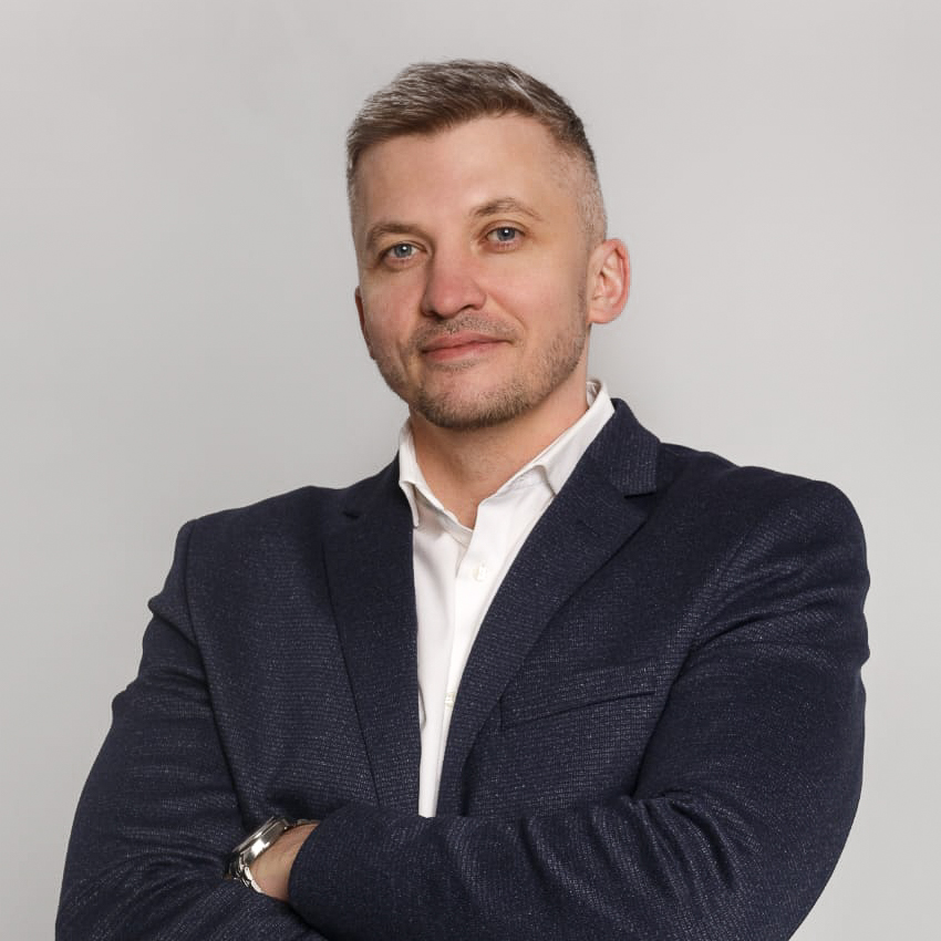 Professionell aussehender Mann mit kurzen, hellen Haaren, der einen marineblauen Blazer und ein weißes Hemd trägt und mit verschränkten Armen vor einem neutralgrauen Hintergrund lächelt.