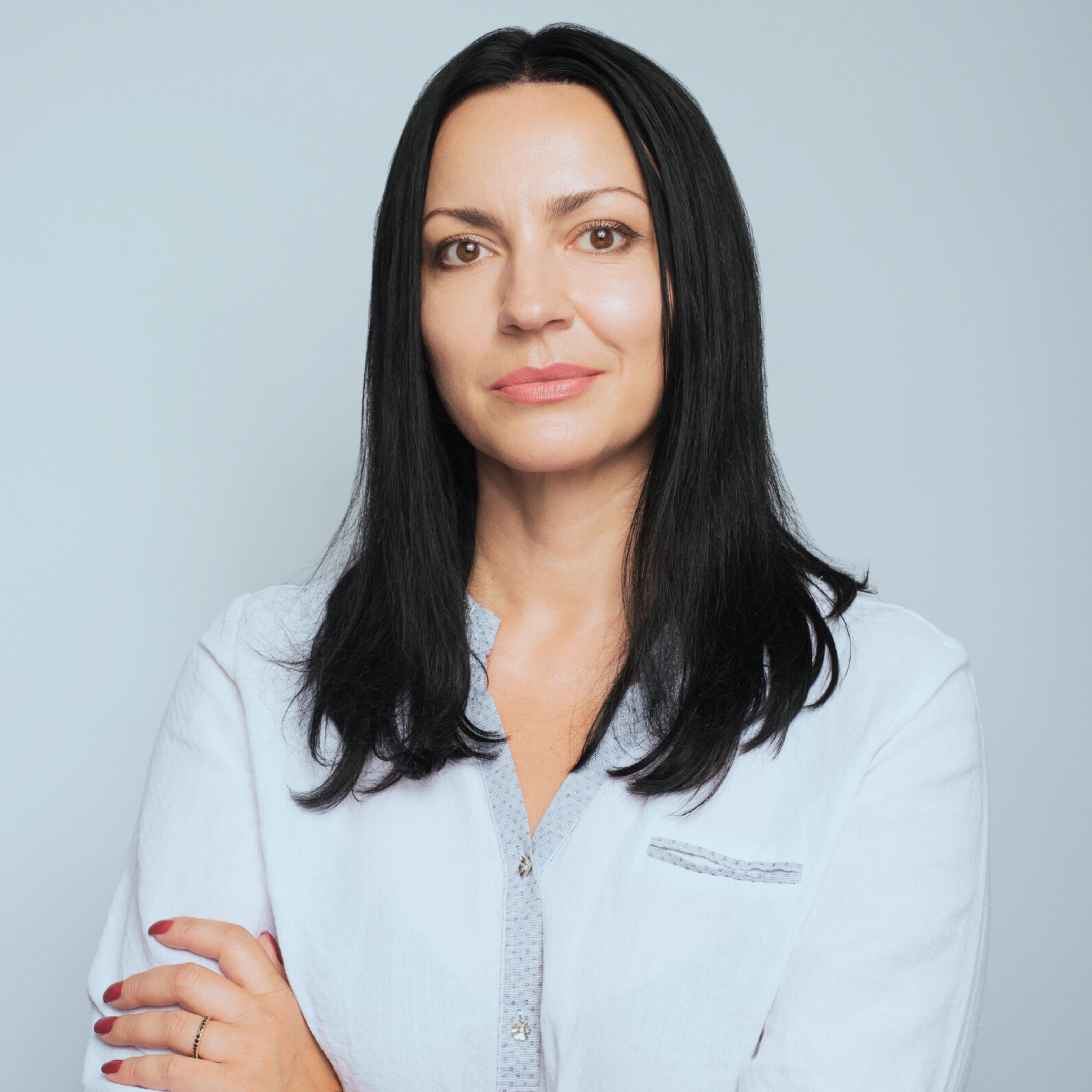 Porträt einer selbstbewussten Frau mit langen schwarzen Haaren, die eine weiße Bluse mit hellgrauem Saum trägt und vor einem neutralen grauen Hintergrund steht.